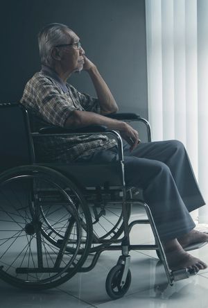 a senior Asian man sitting in wheelchair in a low light room looking contemplatively out a window